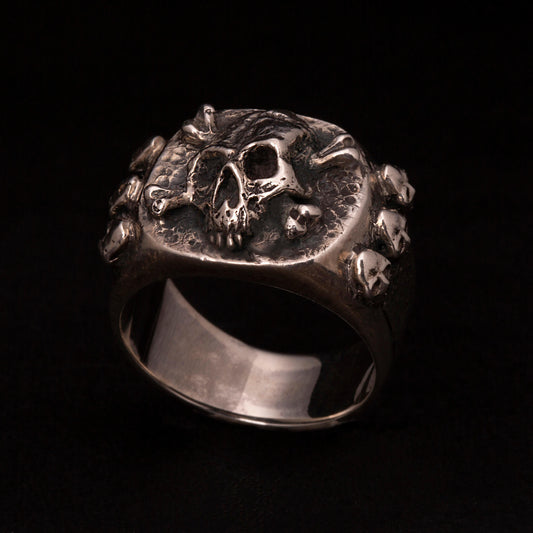 Anello chevalier in argento 925 con teschio e ossa incrociate. Sei micro teschietti impreziosiscono i gambi laterali.