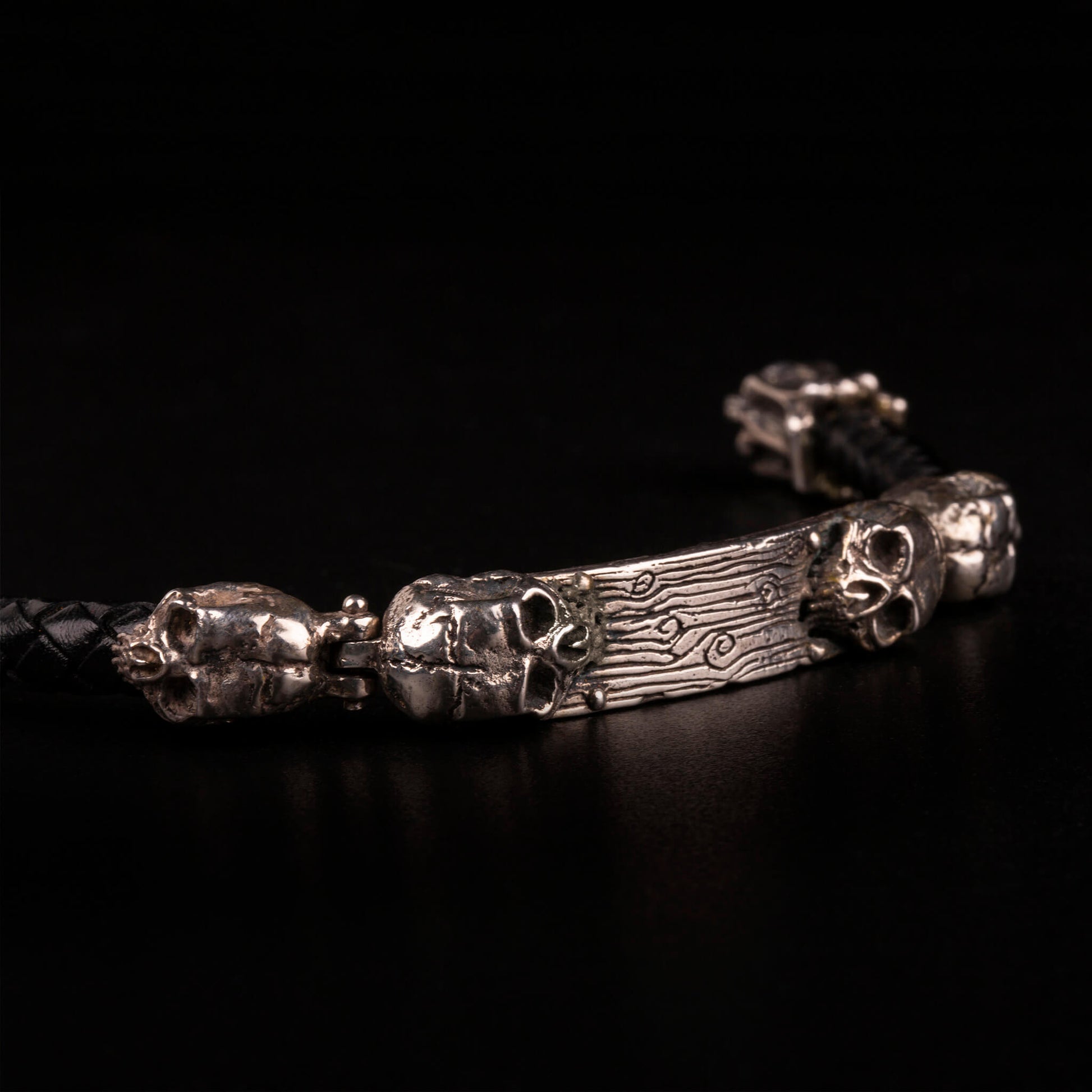 Bracciale con piastrina e teschi ai lati in argento925, cinturino in pelle nera intrecciata e moschettone lavorato in argento925.