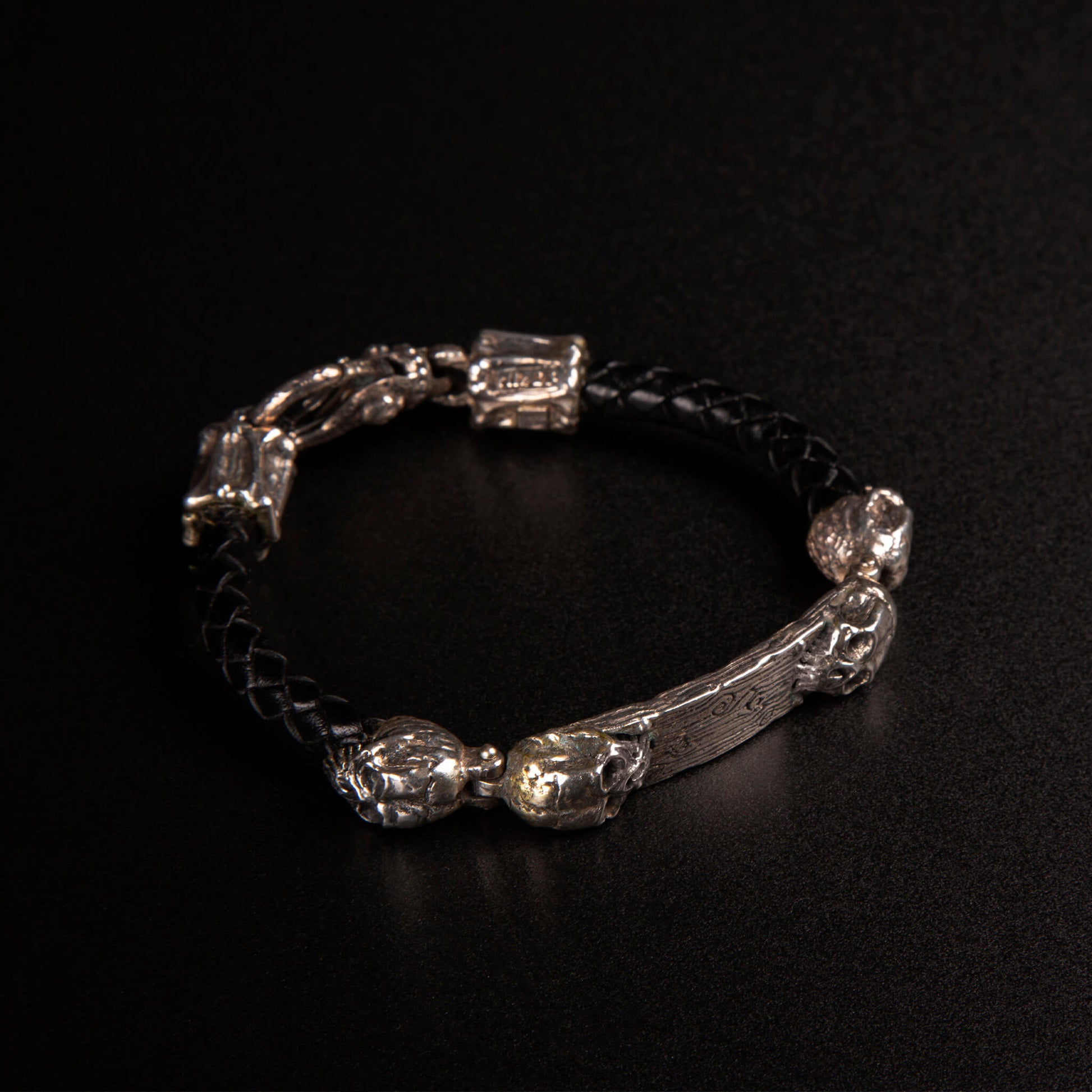 Bracciale con piastrina e teschi ai lati in argento925, cinturino in pelle nera intrecciata e moschettone lavorato in argento925.