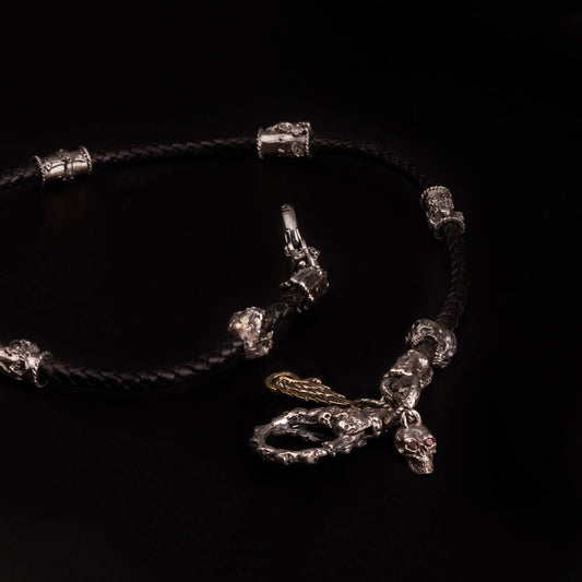 Catena portafoglio con cordino in pelle nera intrecciata ed elementi in argento925 impreziositi con teschi,croci gotiche e cuori votivi. Moschettone impreziosito con piccoli teschi e con targjetta con ala e ruota e teschio con granati.