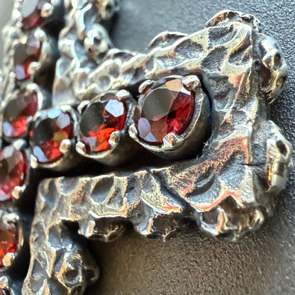 Crux Reliqvia e' un pendente a forma di croce con superficie scalfita irregolare, micro teschi saldati lungo lo spessore e incastonati granati naturali in tutta lunghezza e largezza.